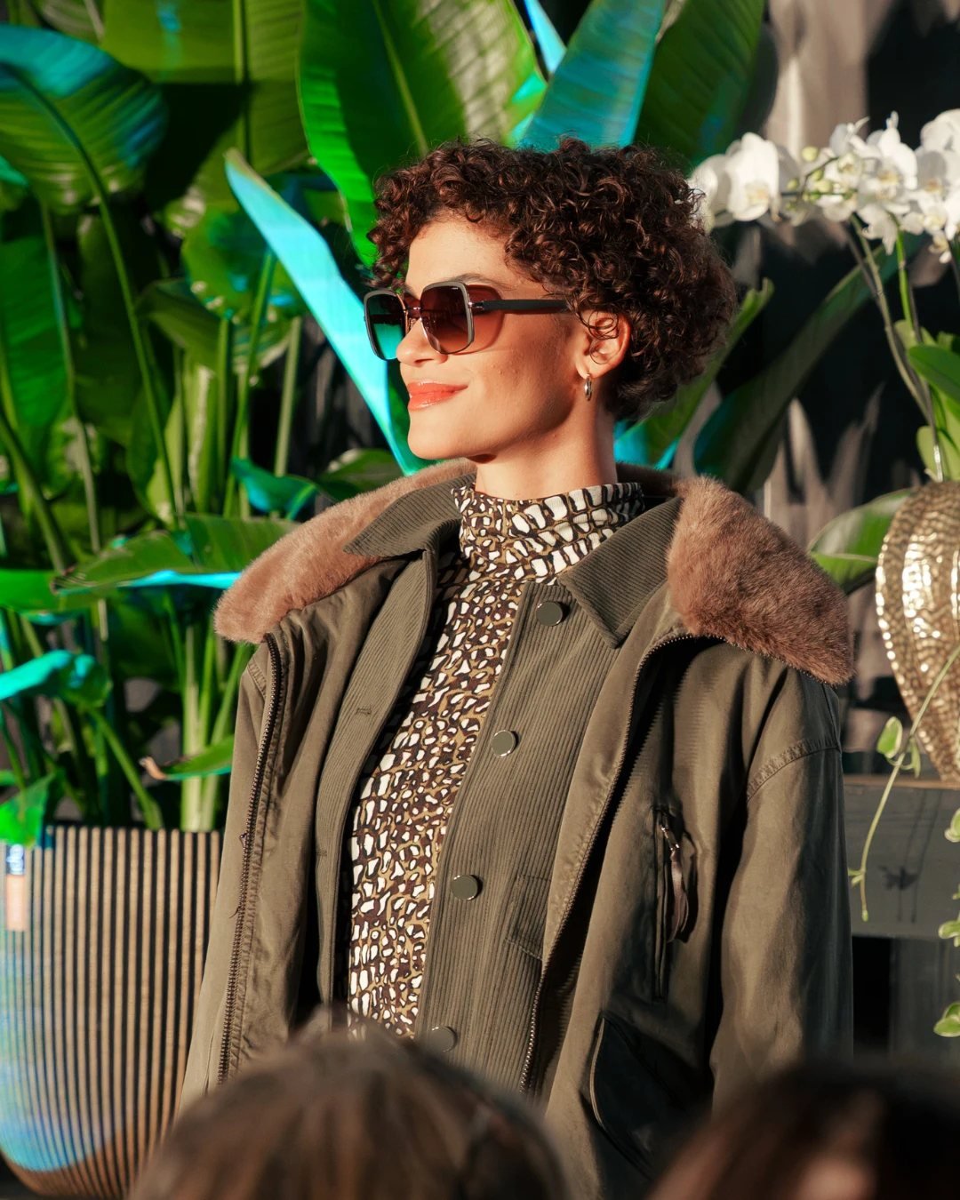 Model mit Sonnenbrille und Cordjacke vor großen Blattpflanzen und Orchideen