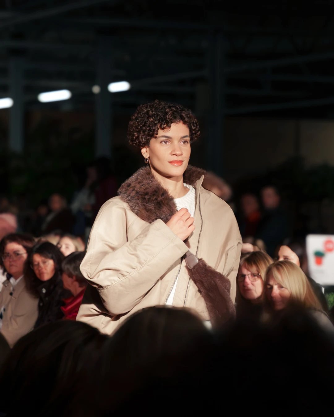 Model in beiger Jacke mit Fellkragen auf dem Laufsteg zwischen den Gästen der Herbstmodenschau